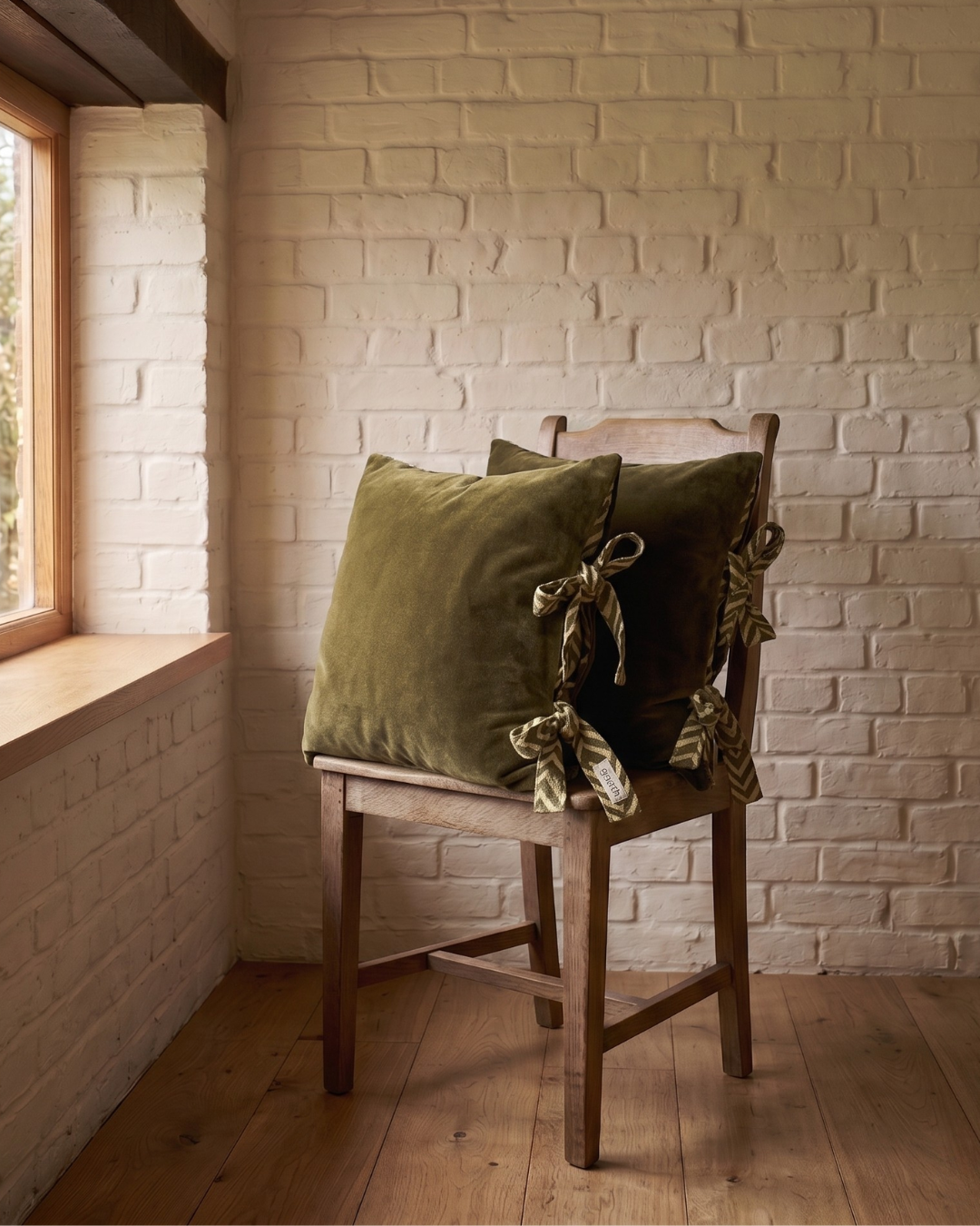 Wooden chair with 2 Moss green Velvet cushions with tie up closure against a white brick wall.