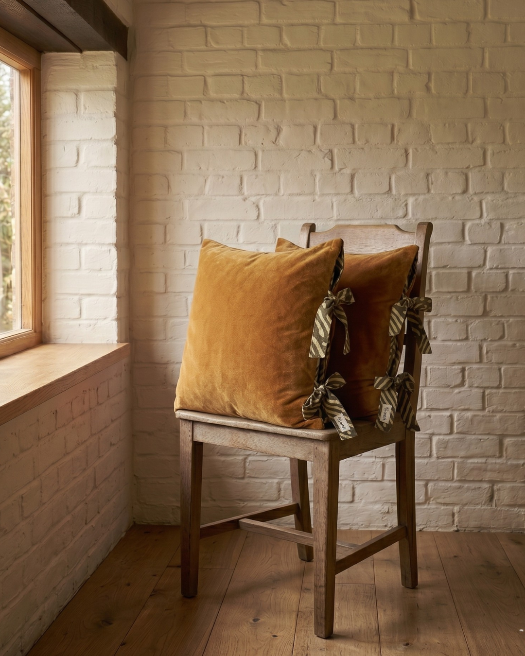 Wooden chair with 2 Ochre brown  Velvet cushions with tie up closure against a white brick wall.